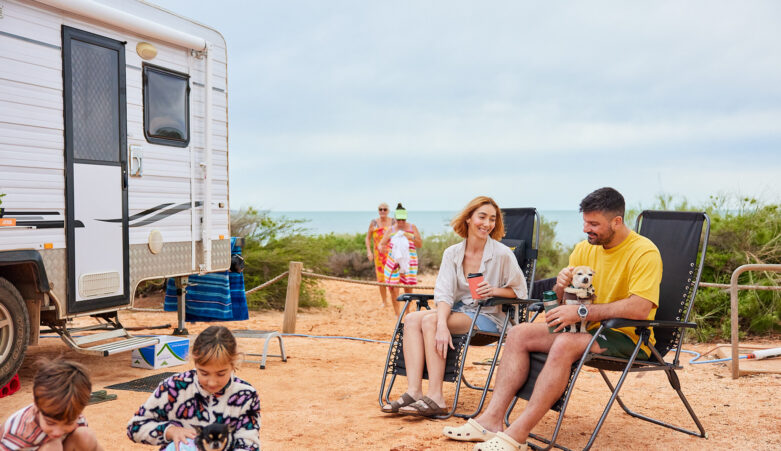 Family of four enjoying beachside campsite, parents sitting on campchairs whilst children play with a dog