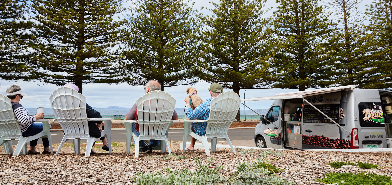 Guests sit at the front of RAC Esperance Holiday Park overlooking the beach and the tall pine trees lining the waterline.
