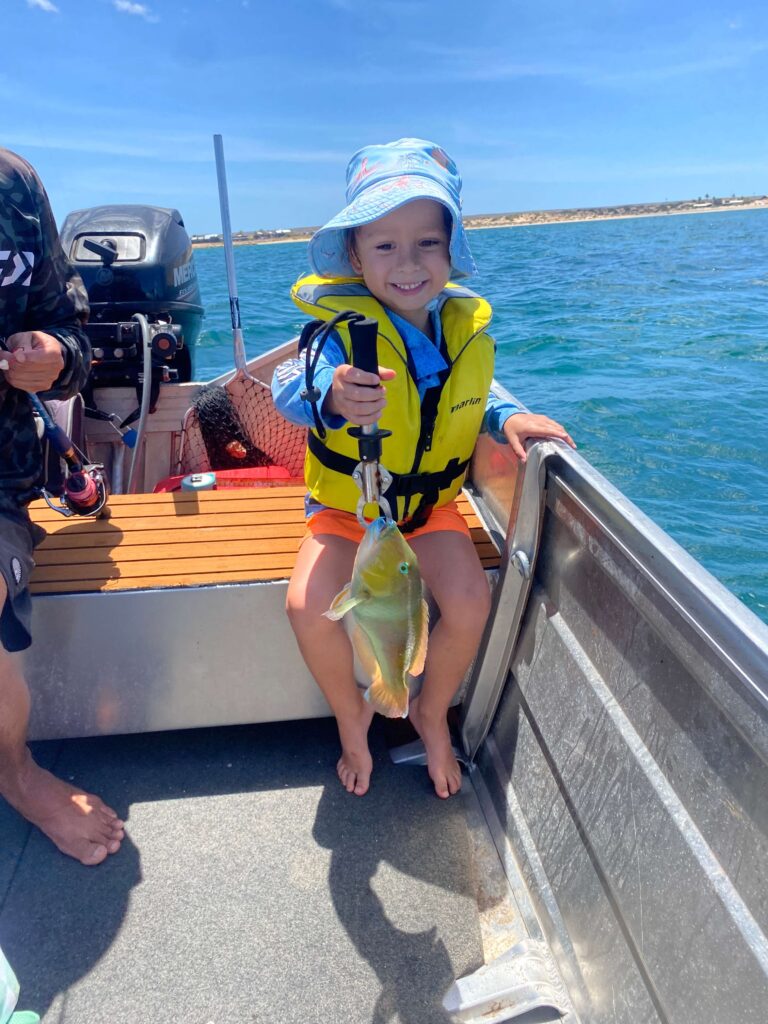 Child with hat and life jacket holding fish on a boat