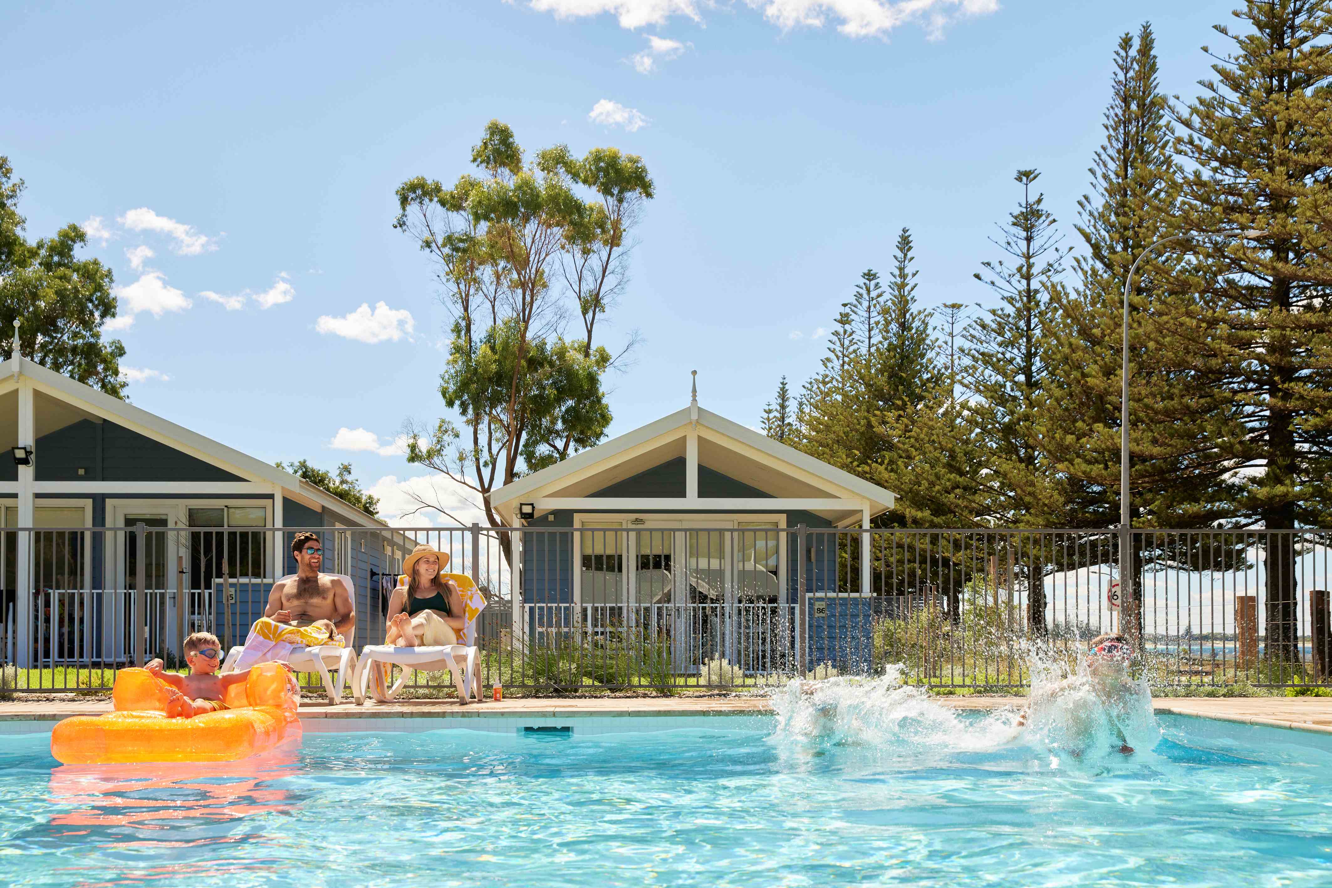 Image of one child making a splash, while their sibling relaxes in a colourful inflatable pool toy. Nearby, parents are relaxing from one of our relaxing pool lounges.