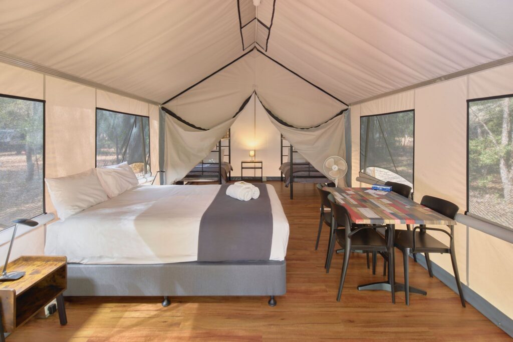 Interior of a Modern Safari Tent, featuring a double bed made with white linen, a table with chairs, and a view through to 2 single bunk beds.